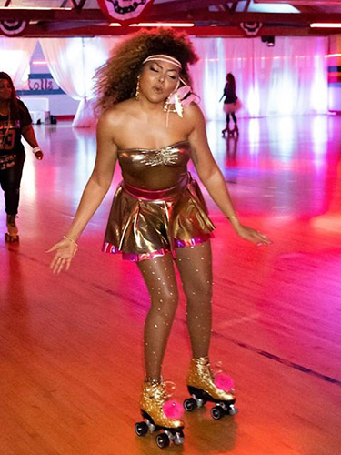 This is a photo of a Black woman, wearin a gold tube top and minny skirt, with pink pieces, and gold skates with pink roses, skating 