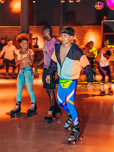  This photo has a White gut wearing blue spandex pants, with white and orange strips. He is wearin a ligth blue, black and neon orange jacket, which is unsipped half way. hea also has a black headband on. There is a ablck couple wearing jeans and tie-die tops, skating behind to him.