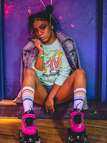 This photo has a Black woman sitting on a purple step, while wearing 9os close. She has on pink glasses; jean jacket; light blue shit with MTV logo on it; white socks with red, orange yellow; and blue stripes on them; and hot pink skates with black laces. She is also holding a lolly-pop.