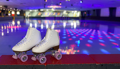 This ia a photo of a par of whit skates resting on a red ledge. The background is of a blurry skating rink with pink and blue tint.